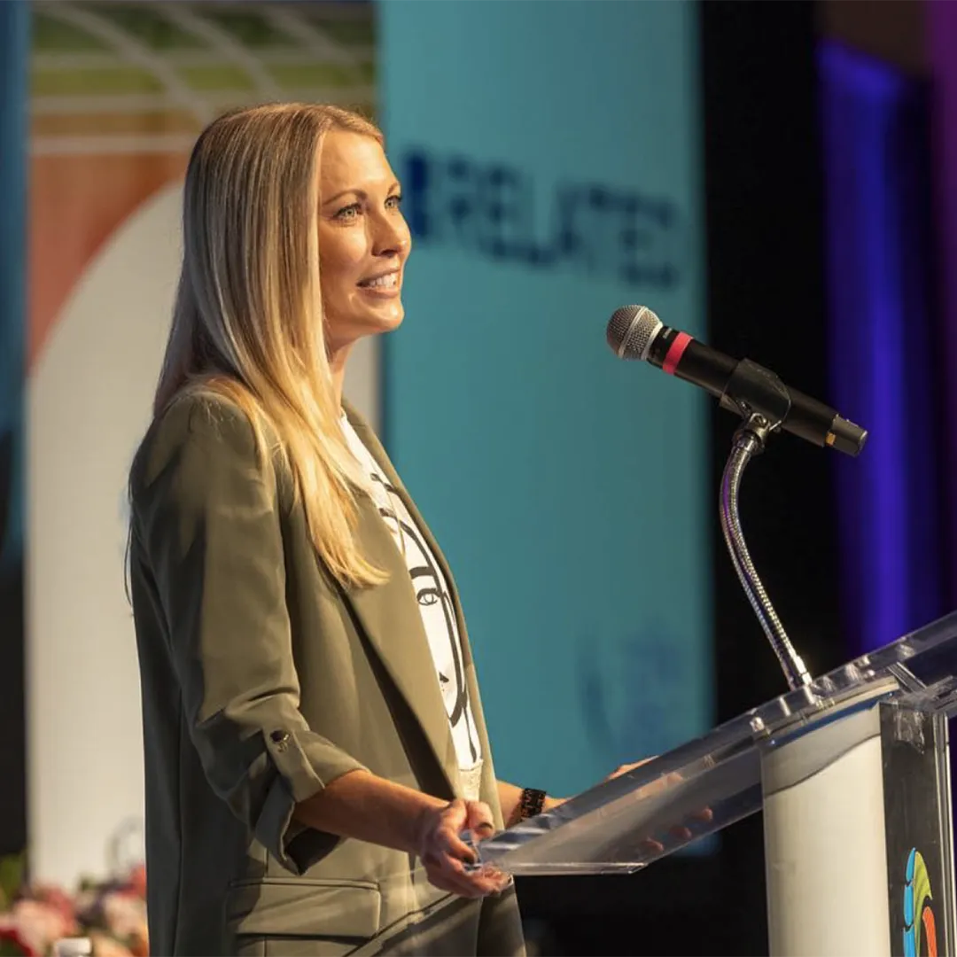 Woman speaks at a podium, addressing an audience with a presentation screen behind her.